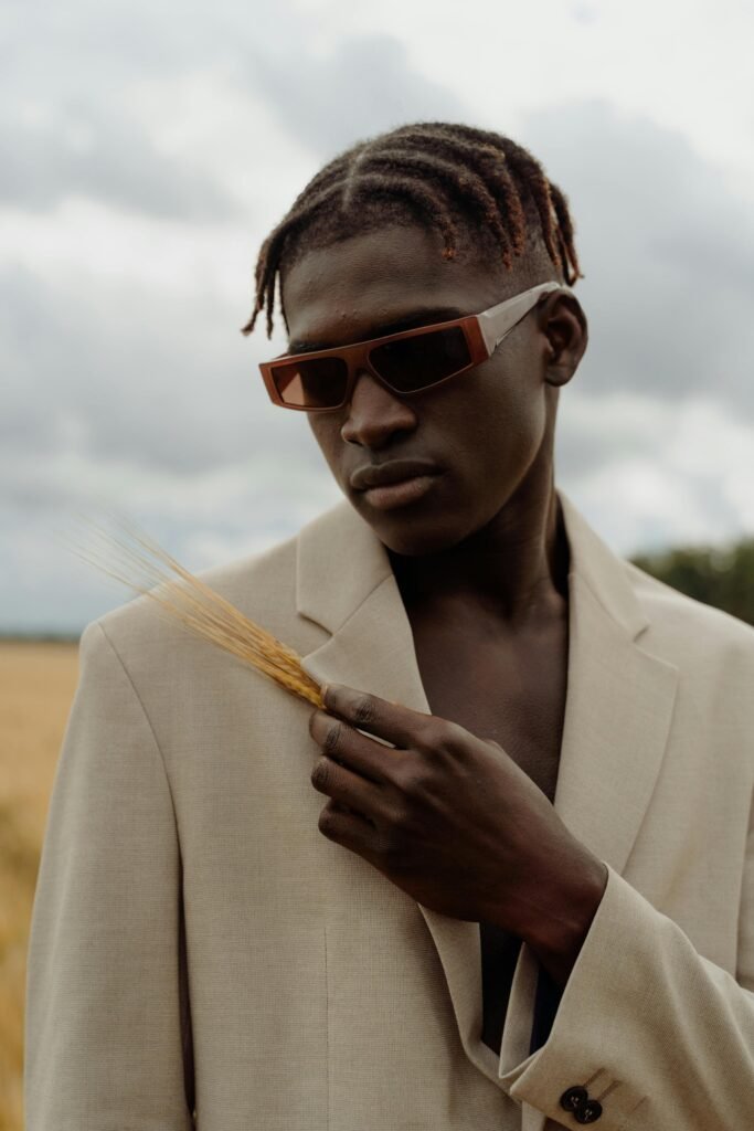 Fashion-forward portrait of a man in sunglasses and a beige suit holding wheat against a moody sky.